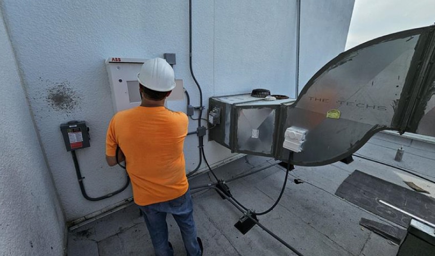A technician in safety gear inspecting rooftop HVAC equipment and an electrical panel.